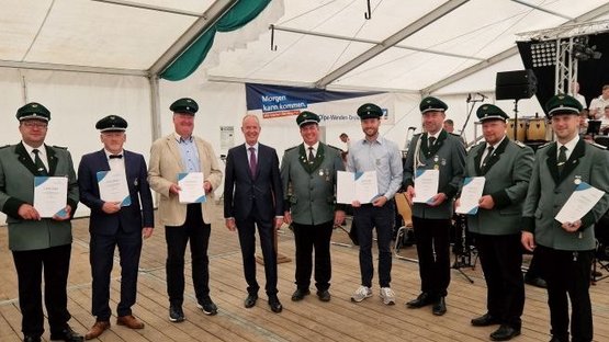 Das Foto zeigt die geehrten Schützenbrüder in grüner Uniform.  Sie stehen im Zelt und haben vom Landrat sowie stellv. Bezirksvorsitzenden Theo Melcher, der in der Bildmitte steht, gerade ihre Urkunde vom Volksbund erhalten.