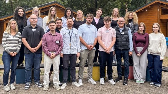 Das Foto zeigt den Regierungspräsident Heinrich Böckelühr (vierte Person von rechts) mit der Ausbildungsleitung und den 16 Auszubildenden vor zwei der typischen Blockhütten in Ysselsteyn, in der die Gruppen übernachten. 