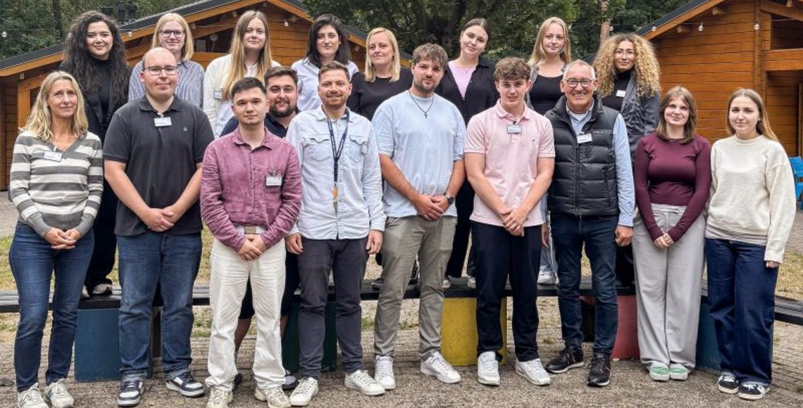 Das Foto zeigt den Regierungspräsident Heinrich Böckelühr (vierte Person von rechts) mit der Ausbildungsleitung und den 16 Auszubildenden vor zwei der typischen Blockhütten in Ysselsteyn, in der die Gruppen übernachten.