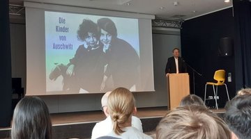 Auif dem Bild sieht man rechts im Bild Alwin Meyer an einem Redepult, hinter him ein großes Bild mit zwei Kinderportraits und der Aufschrift "Die Kinder von Auschwitz", im Vordergrund die Hinterköpfe einiger Schülerinnen und Schüler.
